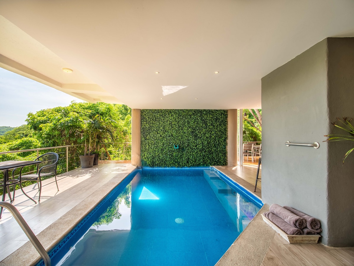 An indoor pool area is highlighted, featuring clear blue water under natural light. Green foliage creates a calming backdrop on one wall. A set of towels is neatly arranged beside the pool, while outdoor seating is visible through an open space, enhancing the relaxed atmosphere.