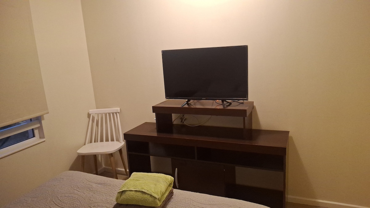 A compact room features a spacious bed with a light-colored coverlet. A wooden dresser with a flat-screen television is positioned above, while a white chair is tucked beside it. Natural light enters through a window, enhancing the neutral wall tones.