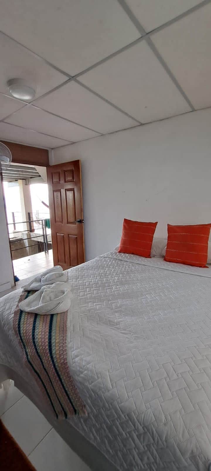 A bright bedroom is featured with a spacious bed dressed in white linens and accented by two orange cushions. A door leads to a balcony area, while soft light filters in through nearby windows, enhancing the room's airy ambiance.