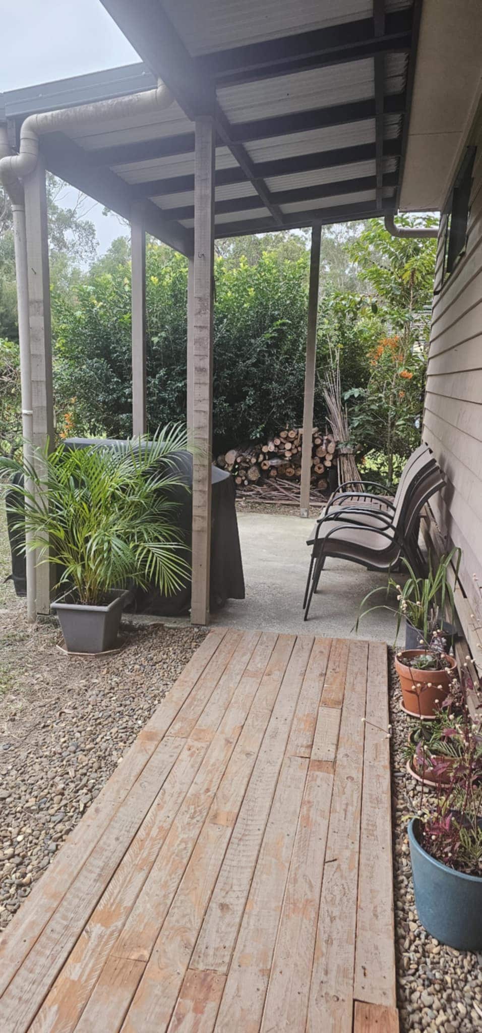 A wooden pathway leads to a covered entryway, framed by potted plants. Sitting chairs are arranged on one side, while a stack of firewood is visible in the background, surrounded by greenery. The atmosphere is calm and inviting.