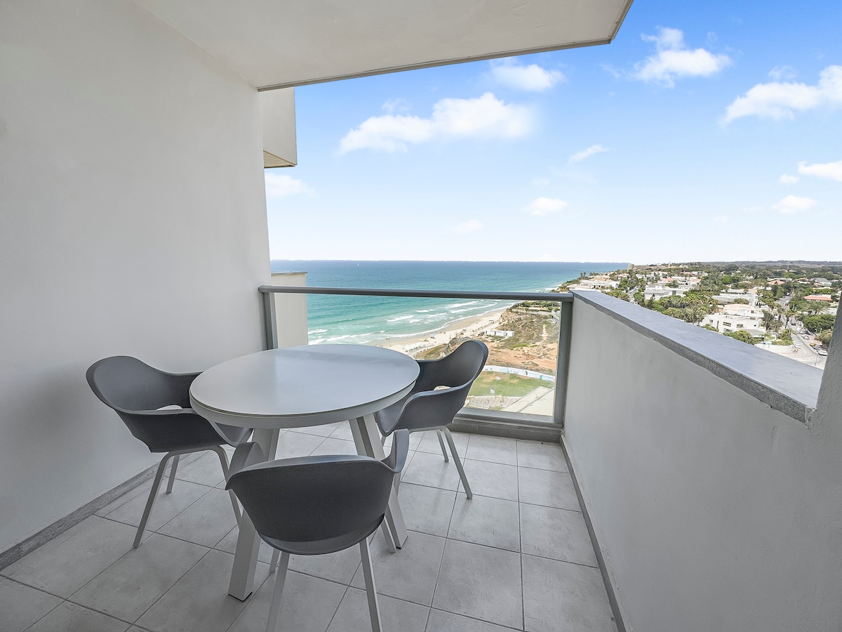 A private balcony features a round table surrounded by four modern chairs, providing a space to enjoy meals or relaxation. The expansive view showcases the Mediterranean Sea and coastline under a clear blue sky.