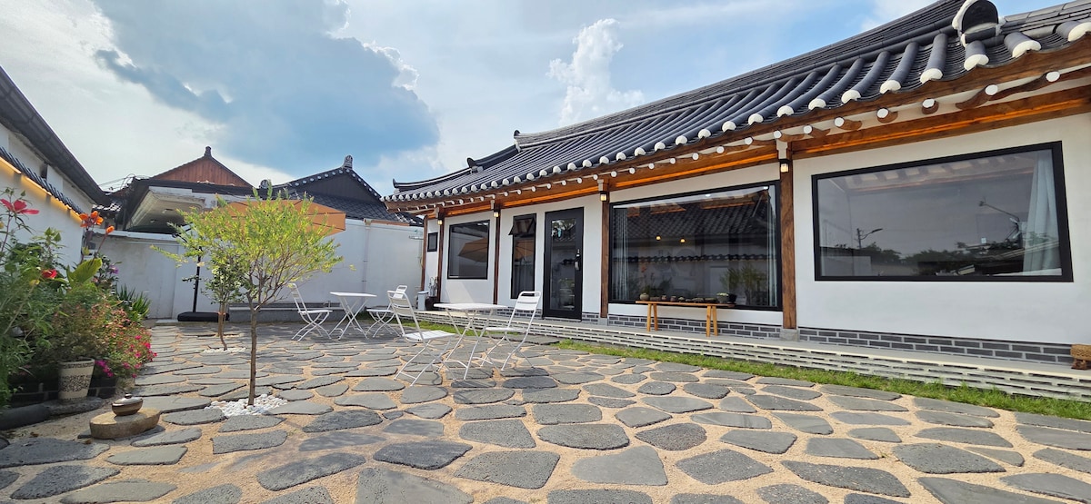 A spacious outdoor area is depicted, featuring a stone pathway surrounded by greenery. A small tree stands in the center, with white chairs and a table arranged nearby. Large windows provide views into the traditional-style building with a sloped roof.