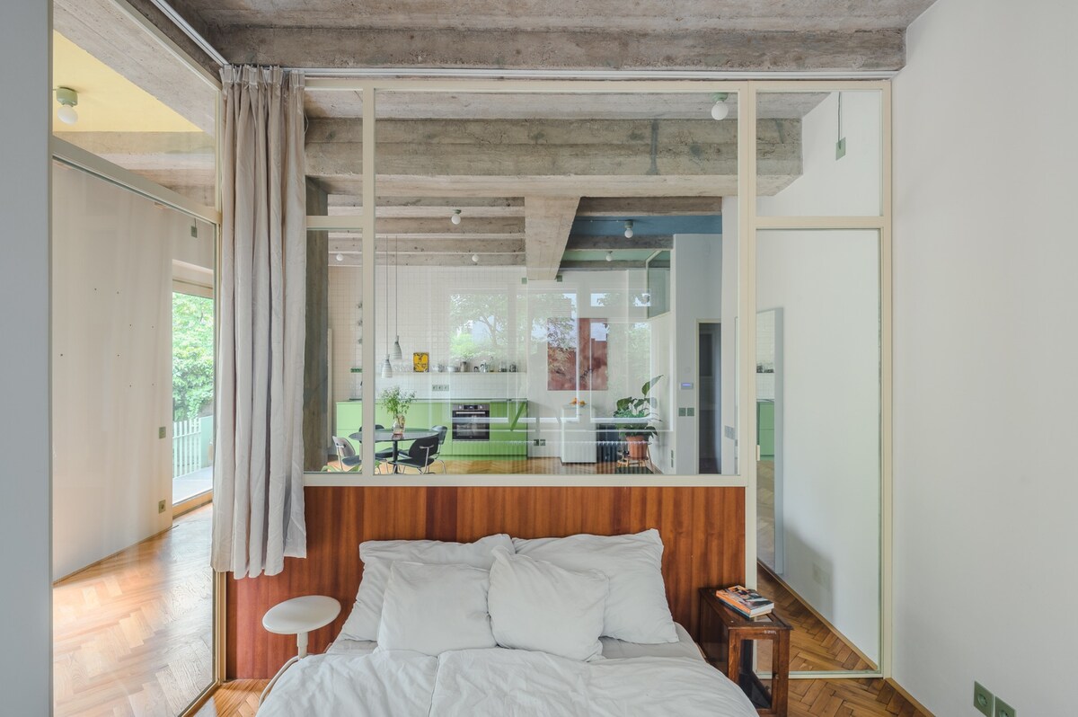 A serene bedroom features a wooden headboard and a spacious bed dressed in white linens. Natural light filters through large glass panels, providing a view of a vibrant living area. Soft curtains frame the window, adding a subtle touch to the calming atmosphere.