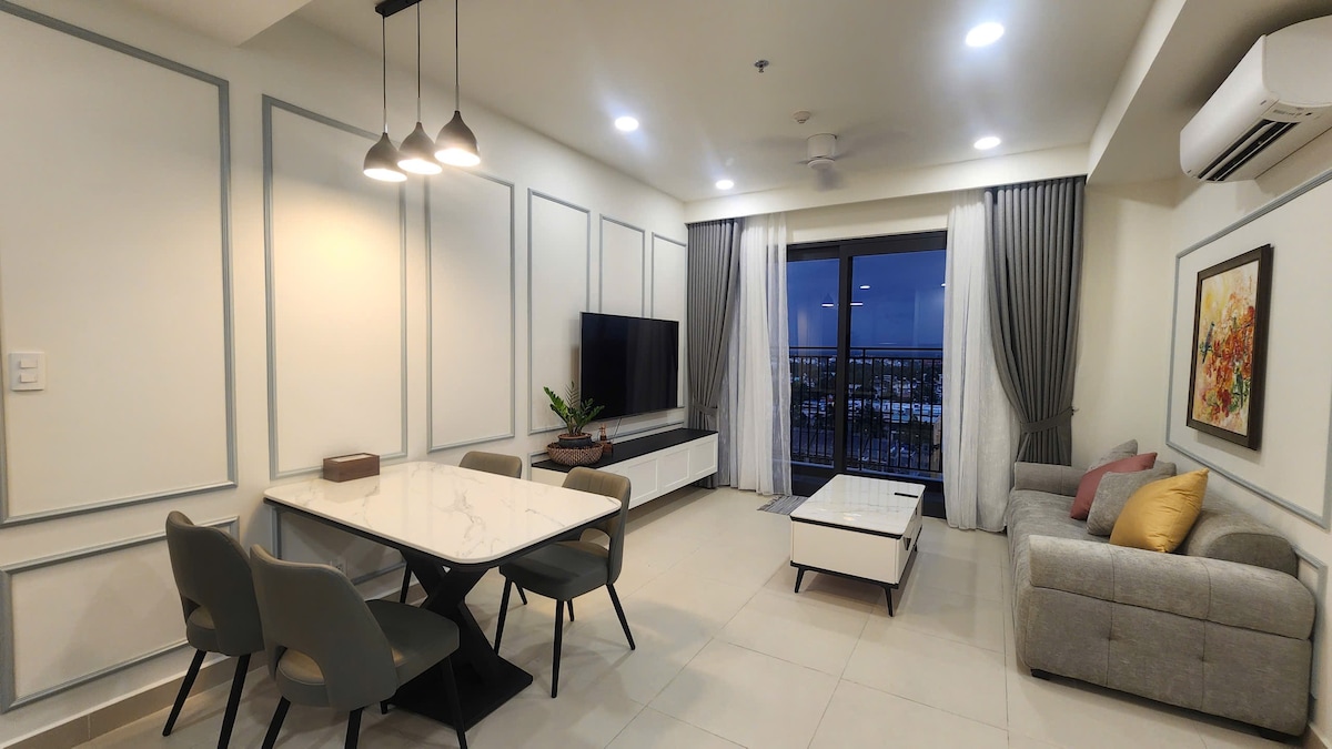 A modern living room features a dining table with four chairs and a comfortable sofa. A large flat-screen television is mounted on the wall, opposite a coffee table. Floor-to-ceiling windows offer views of the outside, with soft curtains framing the scene.
