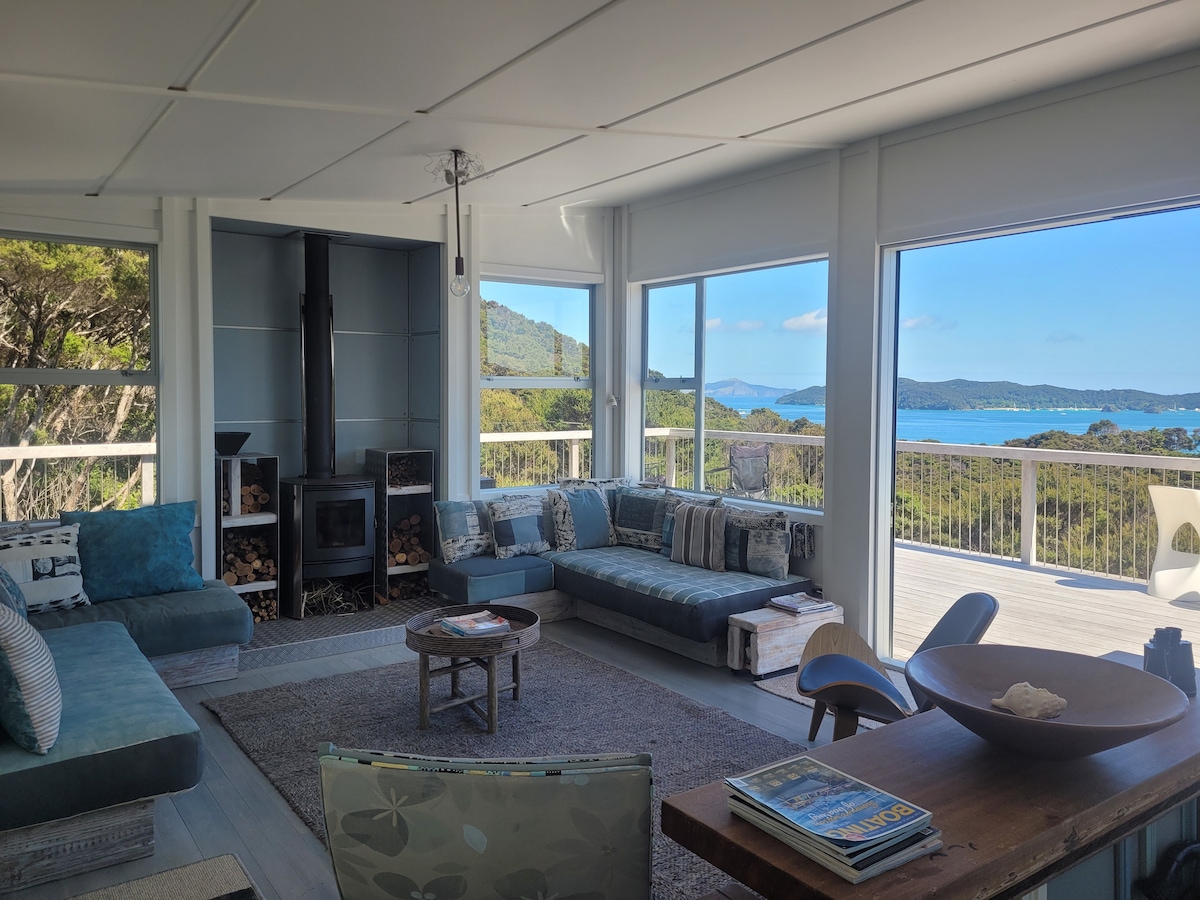 A spacious living area is highlighted by large windows that offer views of the surrounding landscape and bay. Comfortable seating is arranged around a central coffee table. A modern fireplace is situated against a blue accent wall, contributing to the cozy atmosphere.