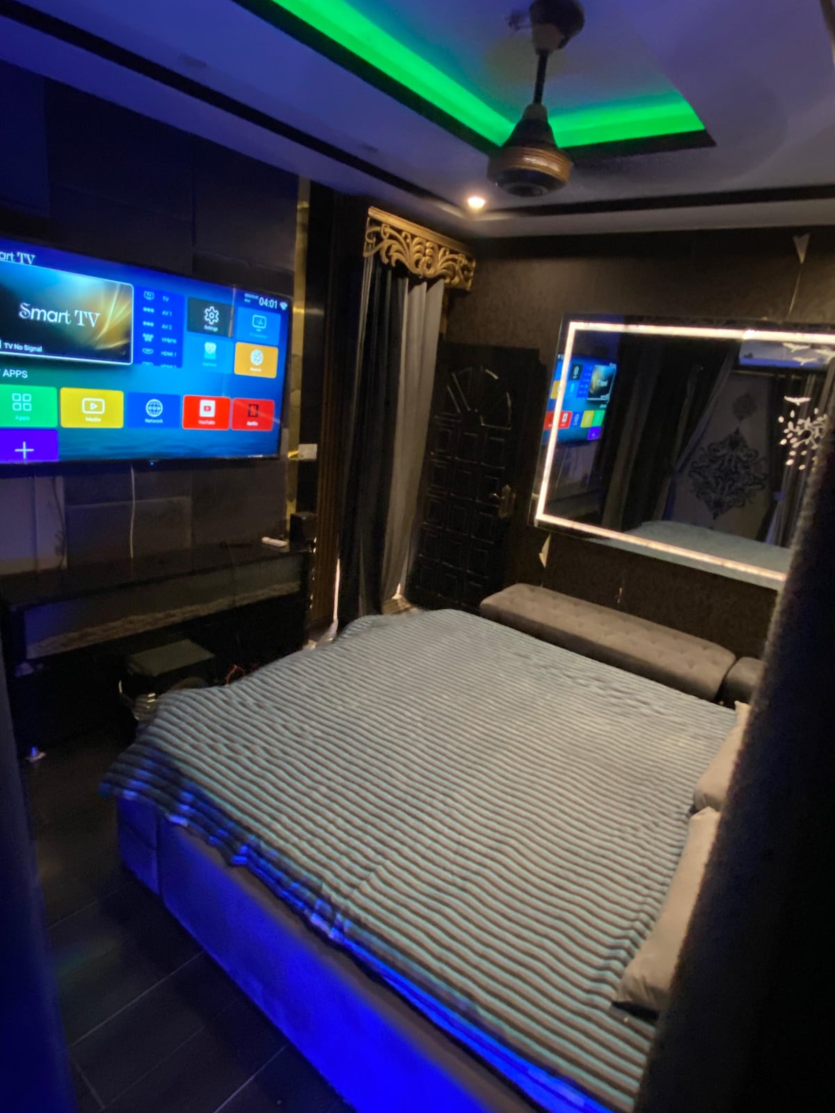 A modern sleeping area features a spacious bed covered with a blue striped blanket. A large mirror and wall-mounted smart TV are positioned above a contemporary cabinet. Soft lighting is provided by an overhead fixture and accentuated by green elements along the ceiling.
