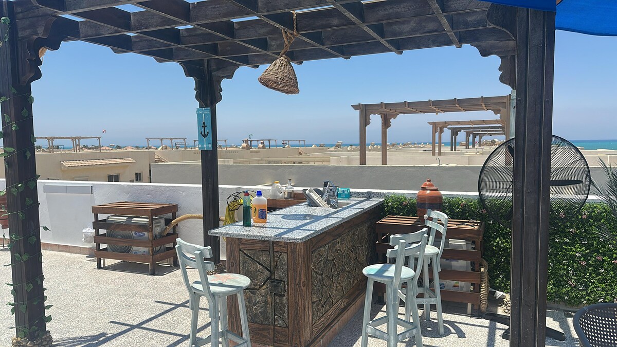 A rooftop terrace is presented featuring a wooden bar with seating for three. The coastal view stretches into the distance, with sun loungers and shaded areas visible. Lush greenery borders the space, adding a touch of nature to the modern design.