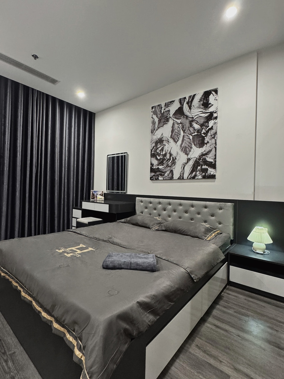 A modern bedroom is presented with a spacious bed featuring a tufted headboard and dark bedding. A soft green lamp sits on a nightstand beside the bed, while a decorative wall art piece adds visual interest above. Dark curtains are drawn to enhance privacy.