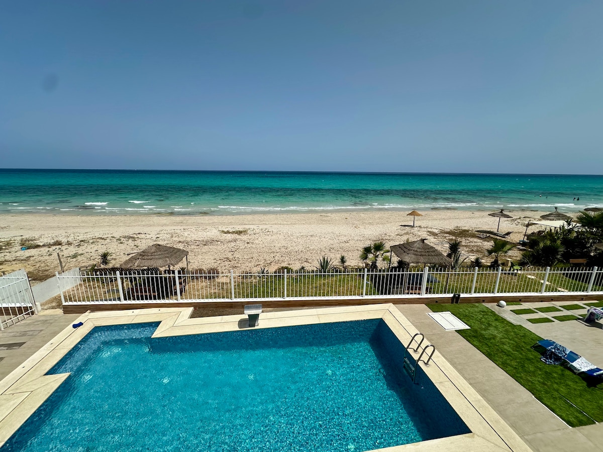 A clear view of a shared pool is presented, with turquoise water reflecting sunlight. The pool area overlooks a sandy beach and calm sea. Several thatched umbrellas are visible on the beach, surrounded by gentle waves and a distant horizon.