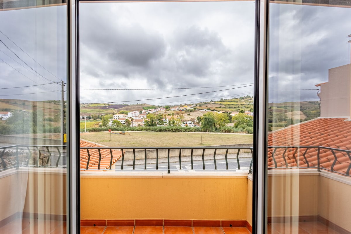 A panoramic view is offered from the balcony, showcasing rolling hills dotted with quaint homes and lush greenery. The scene is framed by floor-to-ceiling glass doors, allowing natural light to fill the room.
