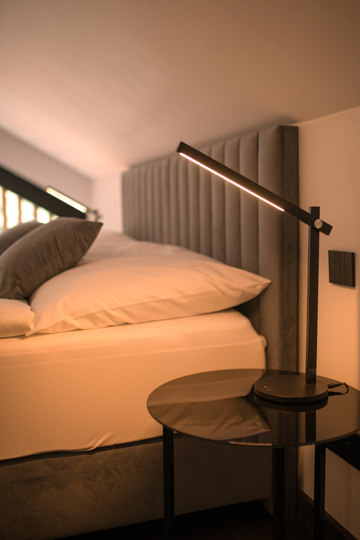 A corner of a serene bedroom features a modern reading lamp beside a spacious bed with layered pillows. A sleek, dark bedside table holds the lamp and provides a minimalist touch. Soft, natural light filters through an angled window, highlighting the textures of the bed linens.