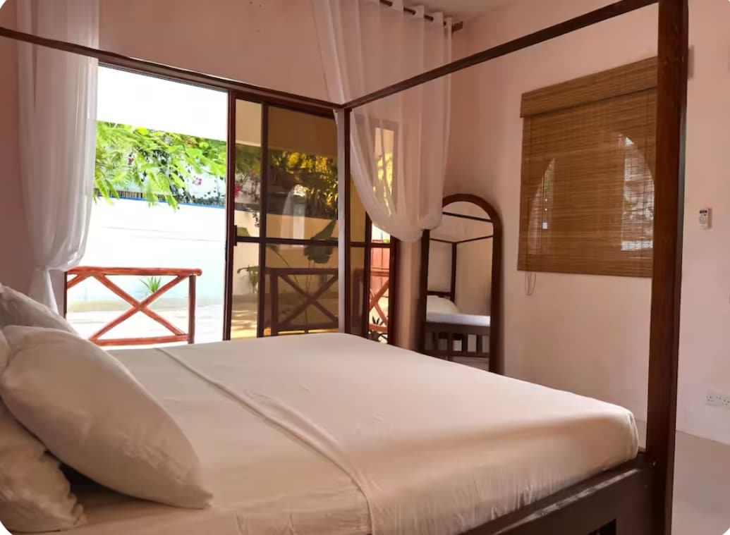 A well-appointed bedroom features a spacious bed with a simple white duvet. Large windows allow natural light to fill the room, while sheer curtains soften the view of the outdoor space. A mirror and a single sofa bed are visible, adding practicality to the design.