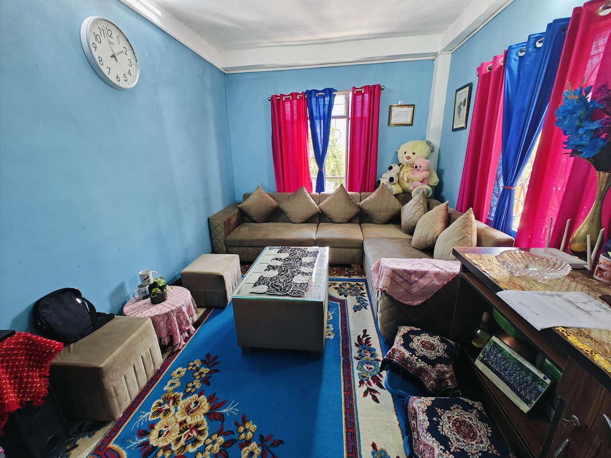 A living room is presented with a sectional sofa arranged around a coffee table. Colorful curtains frame the window, enhancing the blue walls. Decorative items, including plush toys and a large clock, add character. A patterned rug covers the floor, complementing the overall arrangement.