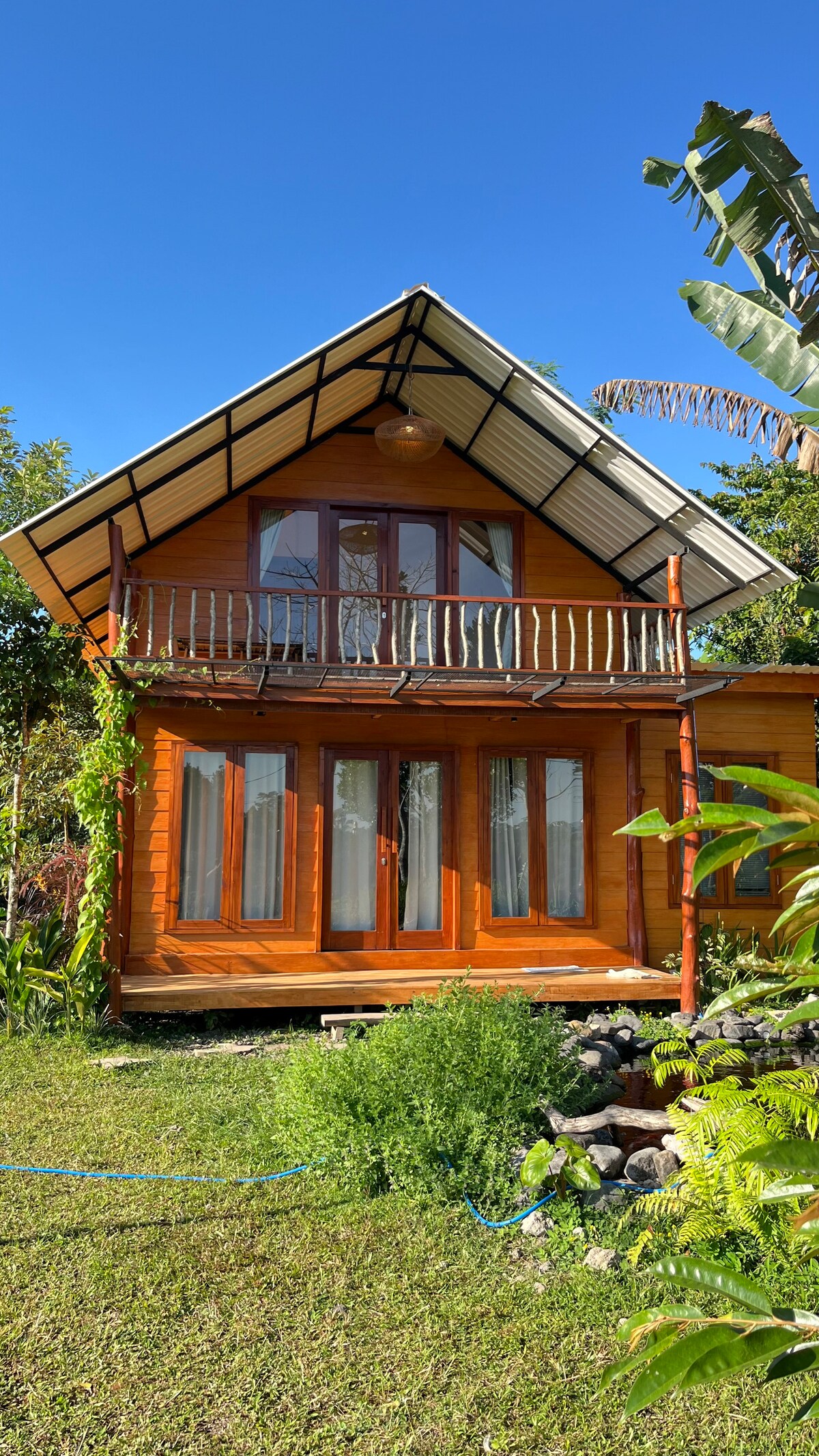 A two-story wooden villa features a sloped roof and a spacious balcony that overlooks the garden. Large windows are present on the lower level, allowing natural light to enter. Lush greenery surrounds the structure, complementing the serene outdoor setting.