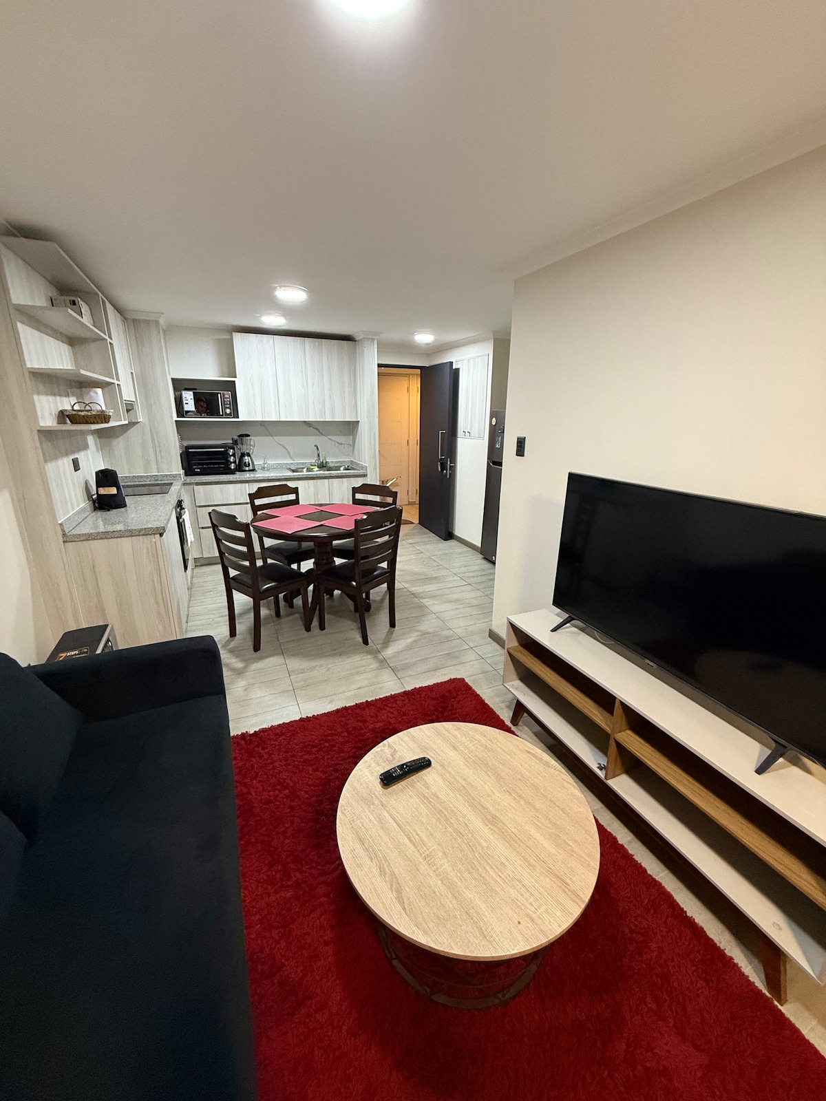 An inviting living area features a dark sofa and a light wooden coffee table resting on a vibrant red rug. A dining table with chairs is visible in the background, alongside a compact kitchen equipped with modern appliances.