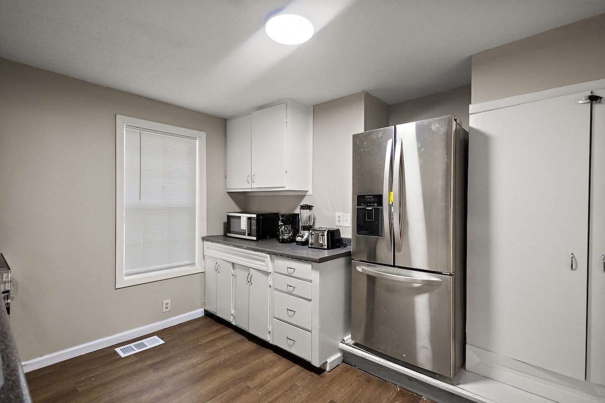 The kitchen features modern stainless steel appliances, including a refrigerator and microwave. A countertop provides preparation space, while cabinets offer storage. Natural light enters through a window, enhancing the room's functionality. A door leads to a pantry or additional storage area.