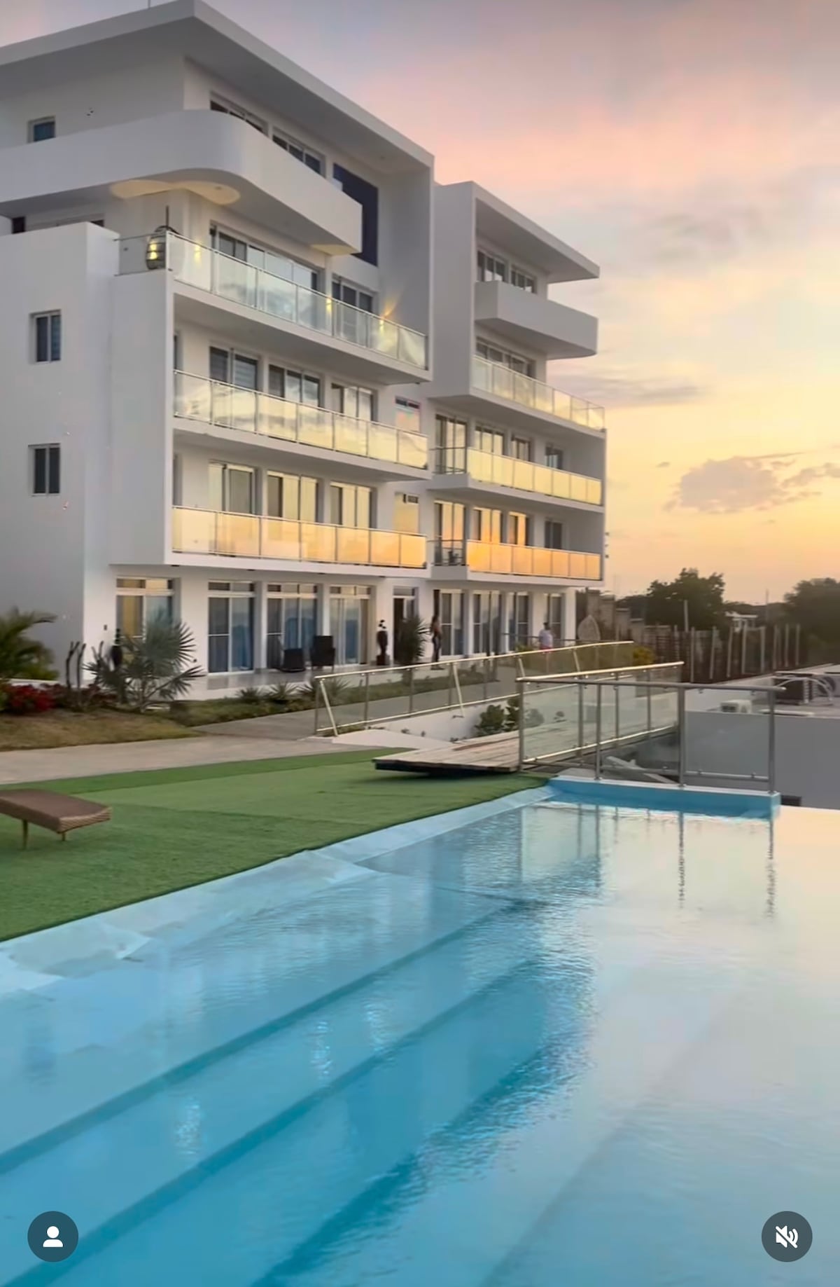 A modern white building is shown against a colorful sunset sky. Glass balconies reflect warm hues, and a tranquil pool with a smooth edge mirrors the architecture. Lush greenery is visible in the foreground, enhancing the serene environment.