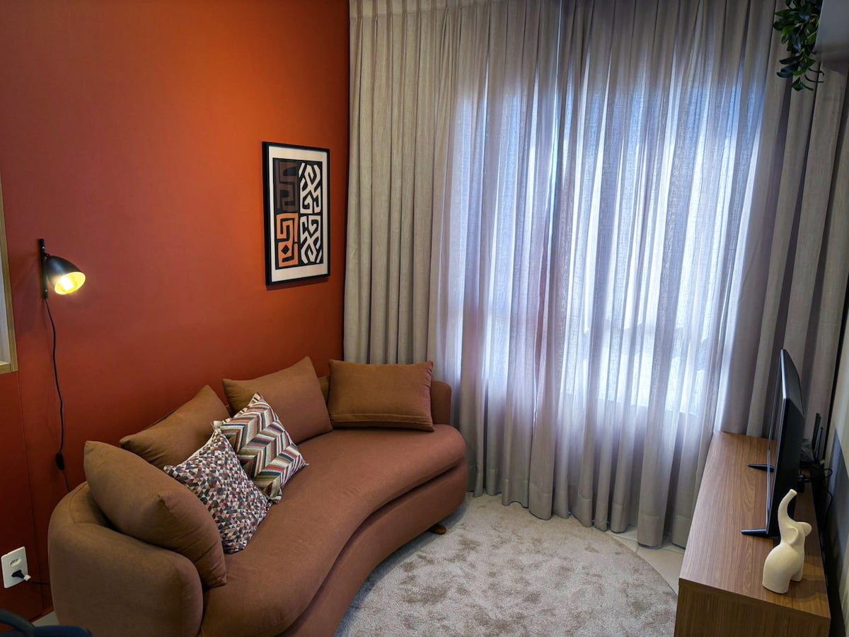 A comfortable seating area is presented, featuring a curved sofa adorned with decorative cushions. A modern artwork is displayed on the wall, complemented by a tall lamp providing soft lighting. Light is filtered through sheer curtains, adding a gentle ambiance to the space.