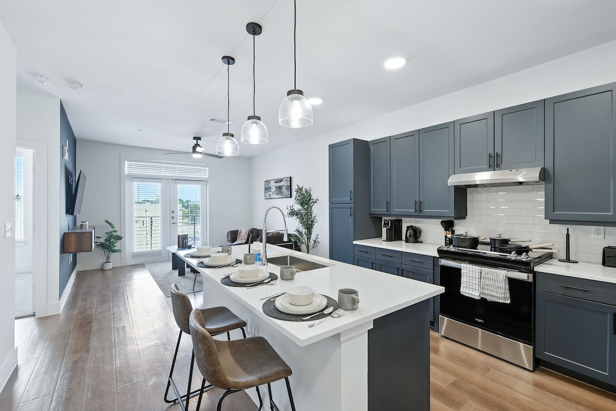 A modern kitchen is featured with dark blue cabinetry and stylish pendant lighting. An expansive island with seating is positioned in the foreground, complemented by neutral tableware. Large windows provide ample natural light, while premium appliances and a sleek backsplash enhance the contemporary design.