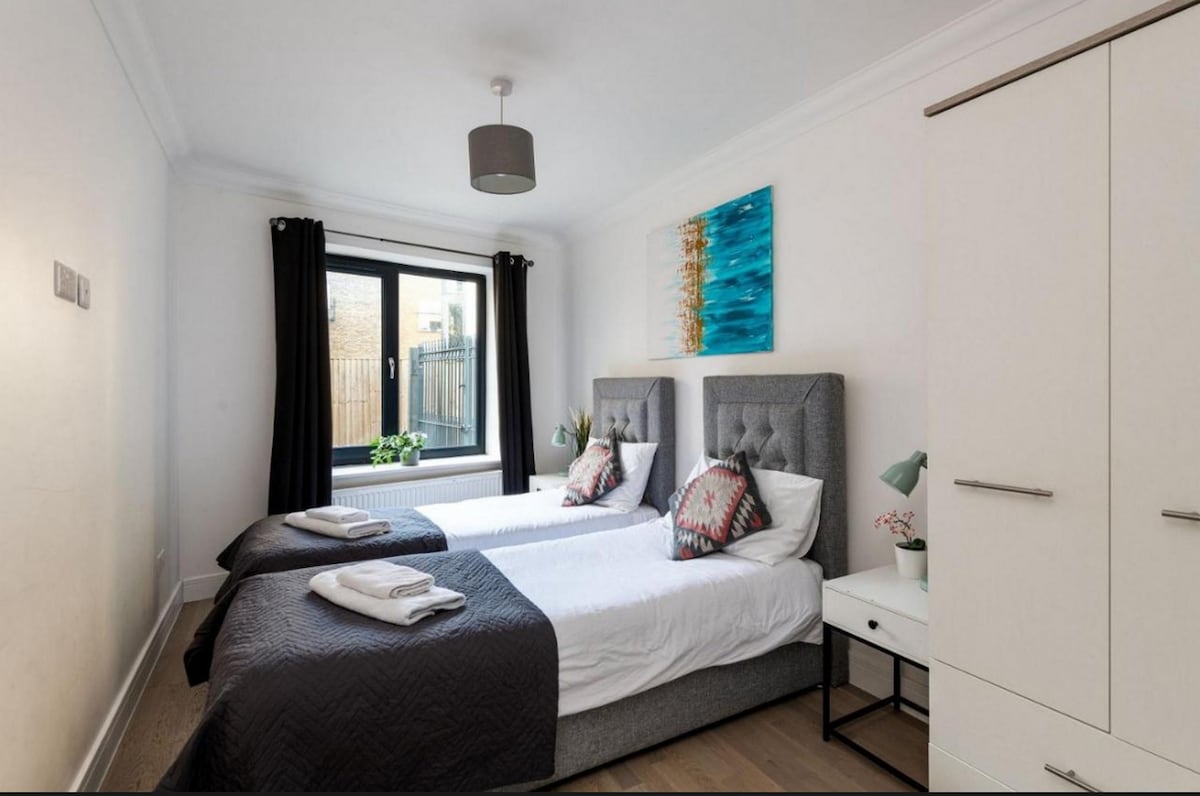 A well-lit bedroom features two single beds with gray upholstered headboards, topped with decorative pillows. A large window allows natural light to fill the room, showcasing a small plant on the sill. A dresser and bedside lamps complement the modern decor.