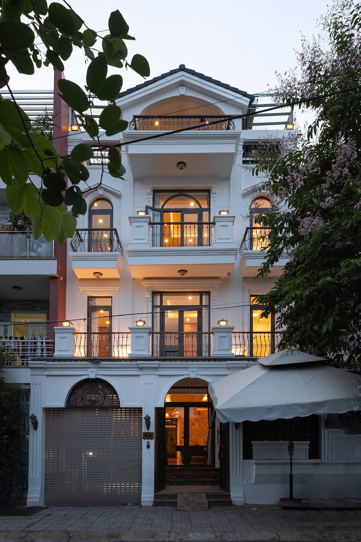 A stylish three-story house is visible, featuring multiple balconies adorned with decorative railings. Warm light glows from the windows, illuminating the facade. Lush greenery and a large umbrella partially frame the entrance, adding to the welcoming appearance of the property.