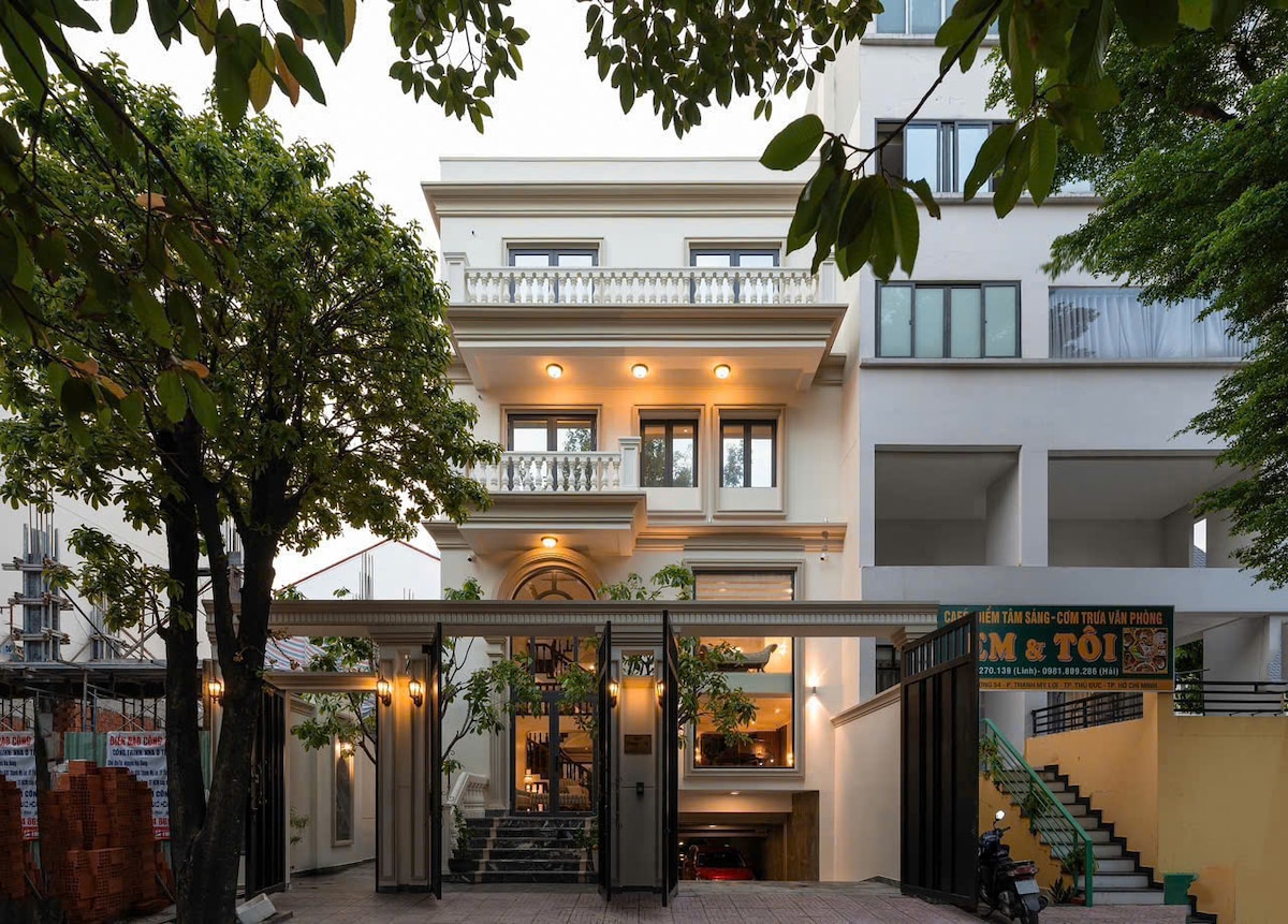 The elegant three-story building features a facade adorned with decorative balconies and large windows. Surrounding trees provide natural shade, while a spacious entrance area is visible, inviting guests into the property. The design complements both modern and classic architectural styles.