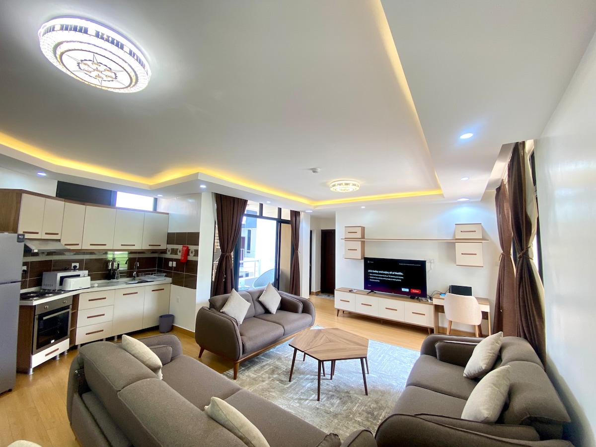 A modern living area is defined by a combination of light wood flooring and elegant finishes. Comfortable seating is arranged around a central coffee table, while a large television is mounted on a wall. Natural light fills the space through expansive windows dressed with dark curtains.