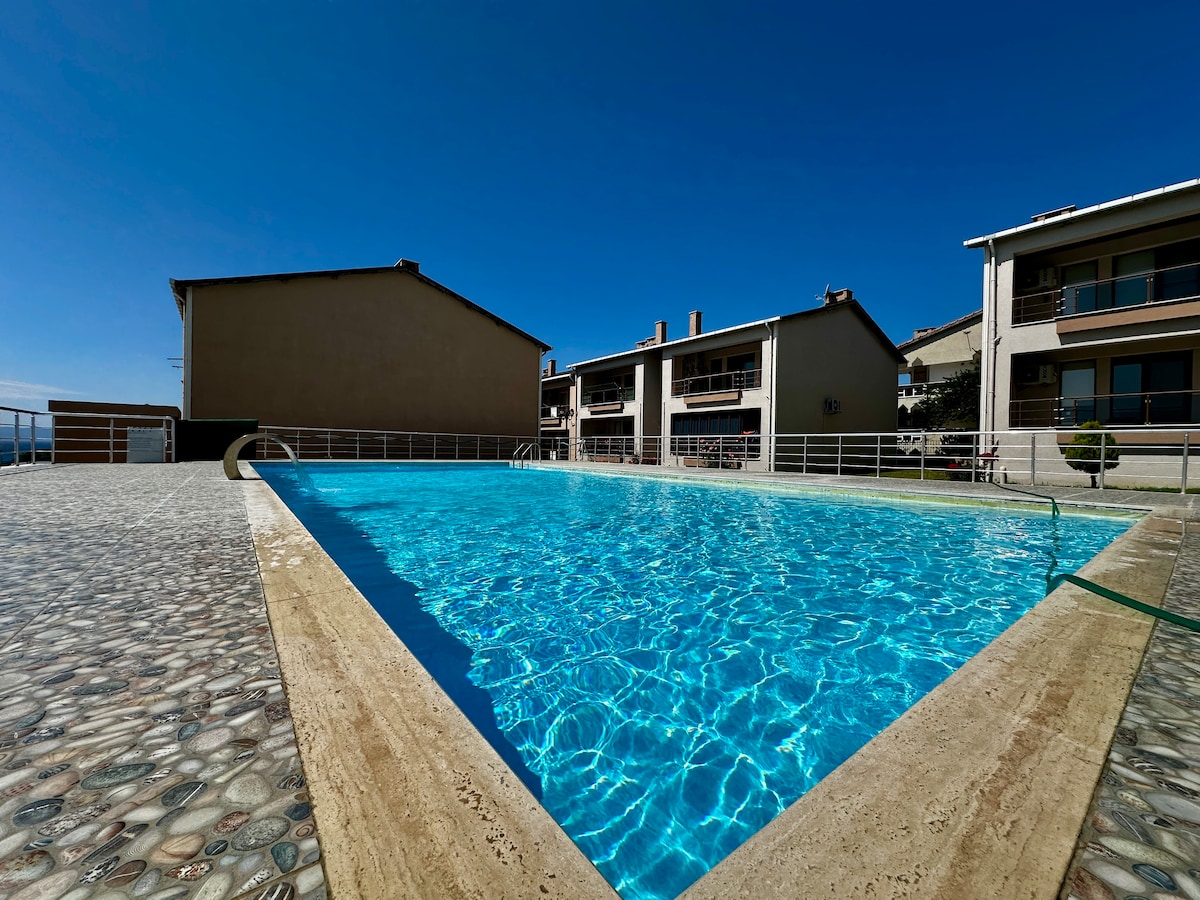 A clear blue swimming pool is set against a backdrop of two multi-story buildings. The pool is surrounded by a spacious deck with a pebbled texture. A bright sky with minimal clouds enhances the outdoor setting.