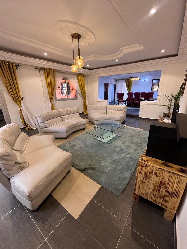 A spacious living area is centered around a glass coffee table, surrounded by two white armchairs and a white sectional sofa. Bright yellow drapes frame the windows, while recessed lighting adds illumination. A textured area rug complements the modern decor.