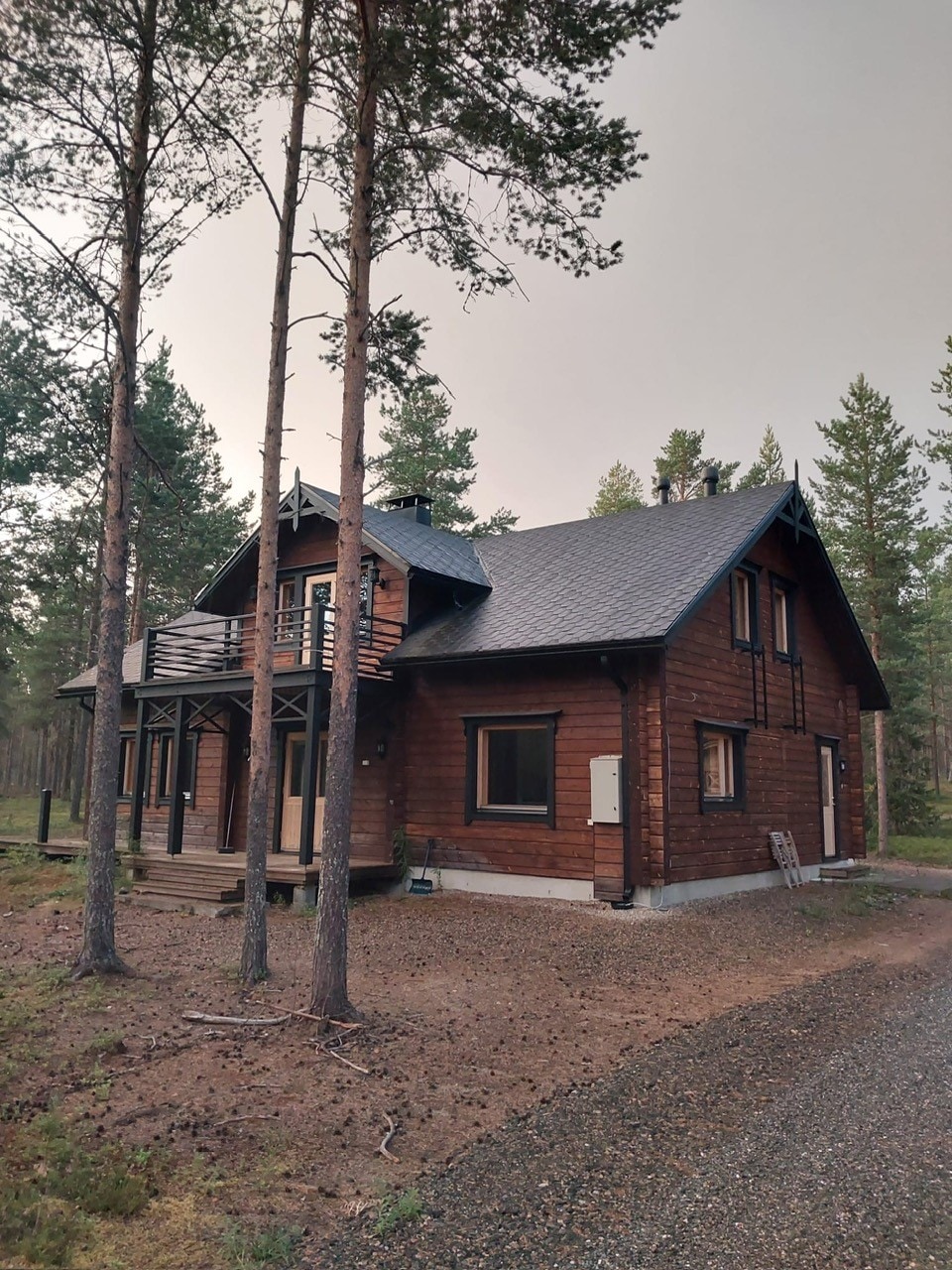 The exterior of a modern wooden cabin is shown, surrounded by tall trees. The structure features a steeply pitched roof, multiple windows, and a small balcony. A gravel path leads to the cabin entrance, blending harmoniously with the natural wooded setting.