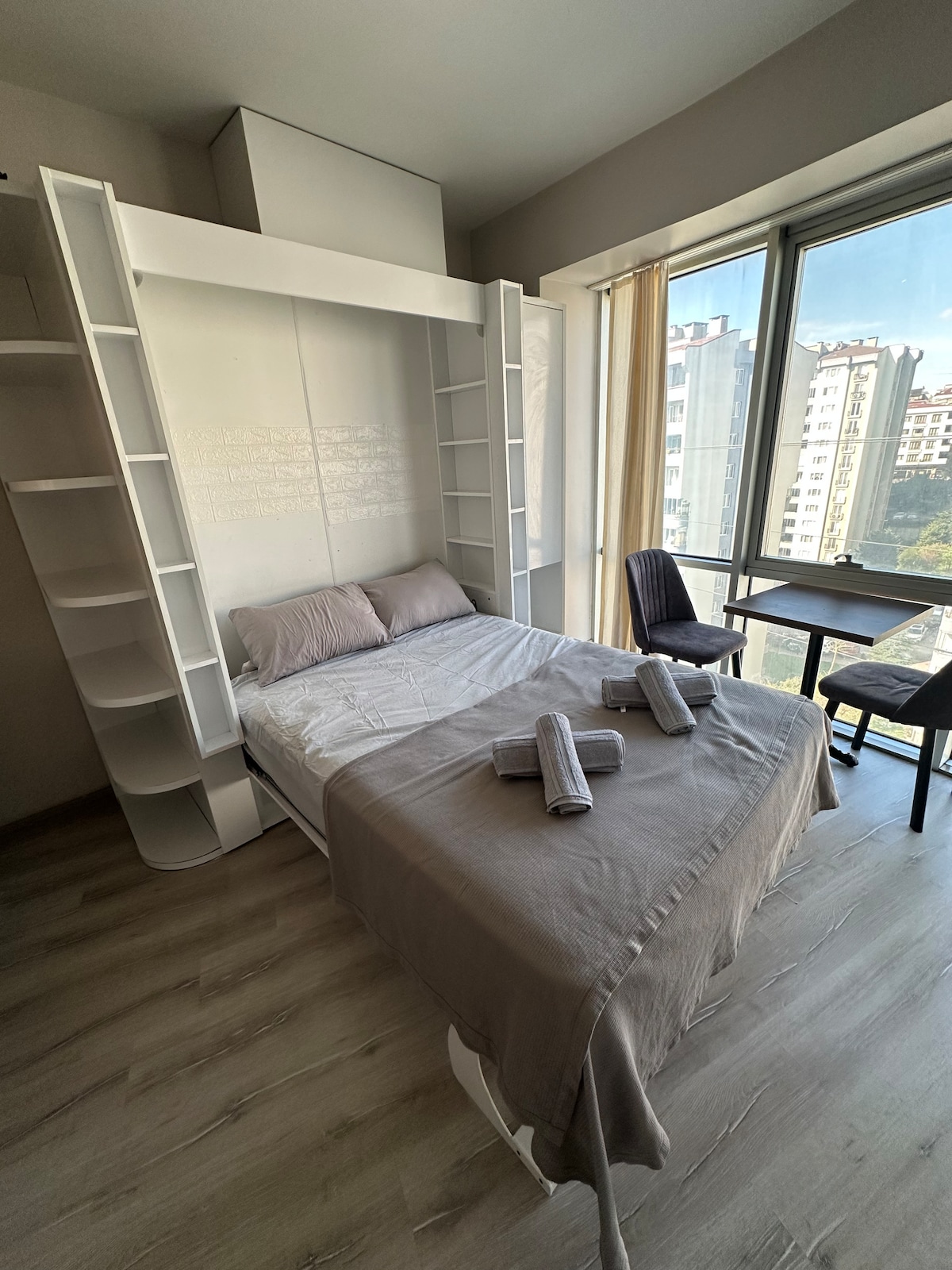 A versatile room features a wall bed framed by white shelving units, complemented by a light-colored quilt. Two rolled towels rest on the bed. A small table and chair are set beside a large window, allowing natural light to brighten the space.