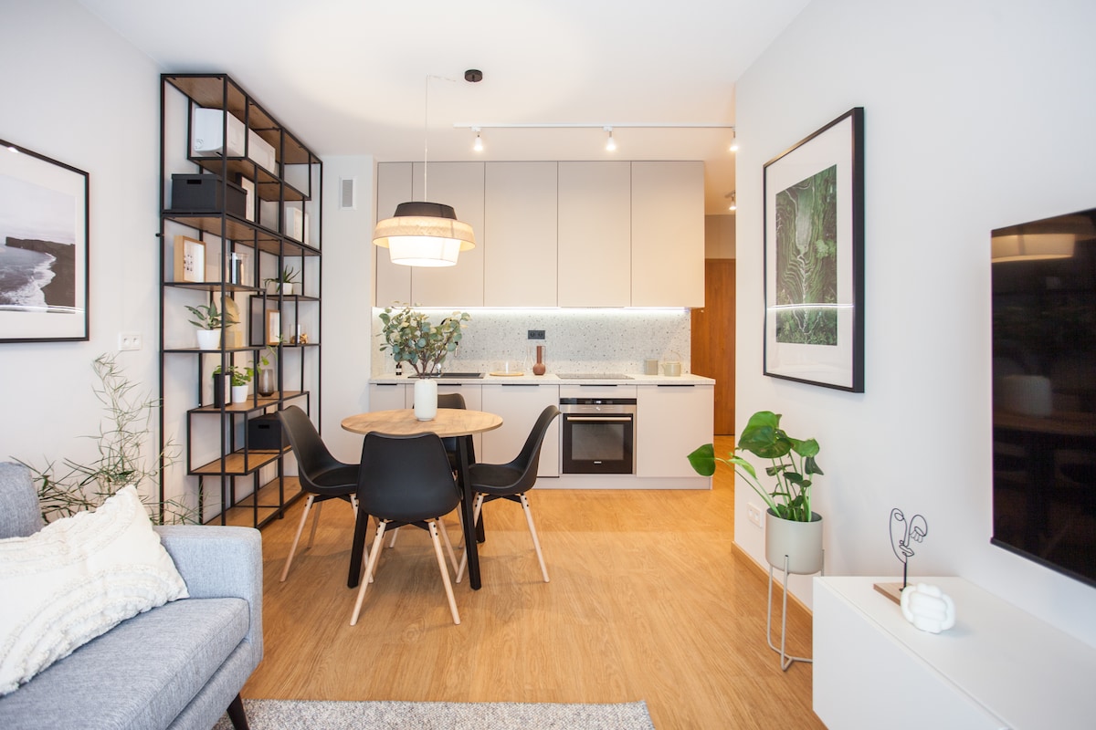 A modern kitchen and dining area feature a round wooden table surrounded by black chairs. The sleek kitchen includes built-in appliances and light cabinets. Greenery from potted plants adds a touch of nature to the space, complemented by soft lighting and neutral decor.