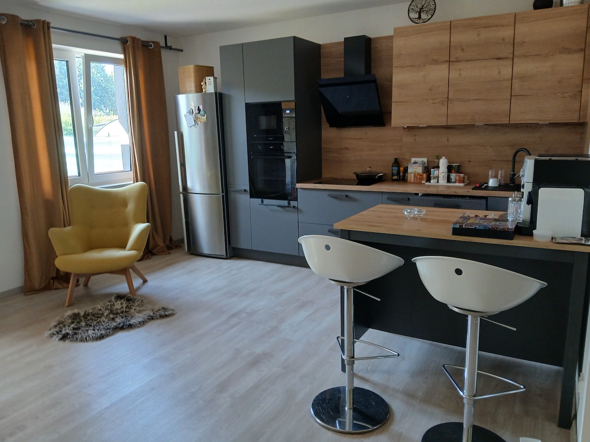A modern kitchen space is characterized by sleek cabinetry and a wooden accent wall. A stylish yellow armchair adds a pop of color near a window. Bar stools are positioned at the kitchen island, while appliances such as a refrigerator and oven are visible in the background.
