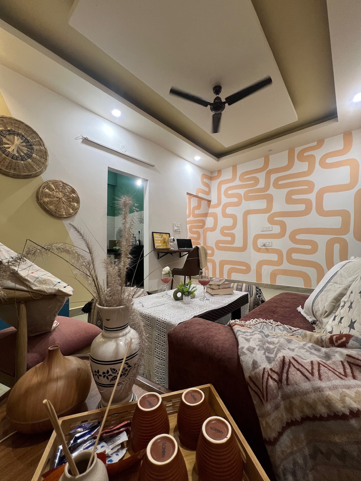 A cozy living area is highlighted by a patterned accent wall in warm tones. A fan hangs from the ceiling, while artful decor, including a decorative tray and vases, enhances the space. A comfortable sofa and textured throw pillows offer inviting seating.