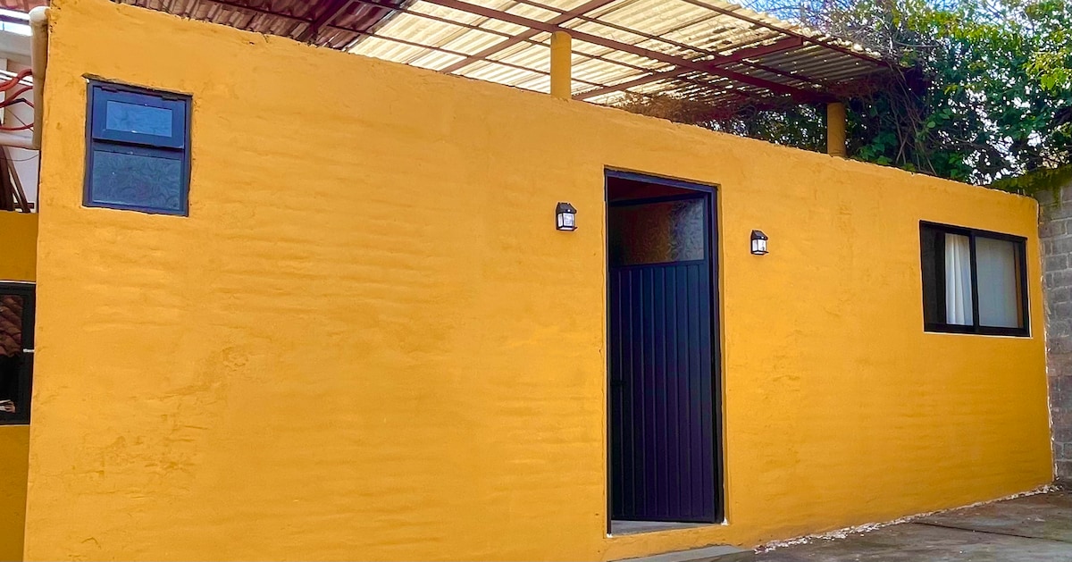 The exterior features a vibrant yellow wall with a textured finish. A dark entry door is centered, flanked by two windows. Overhead, a cover provides shade, while greenery can be seen along the top, contributing to a welcoming environment.
