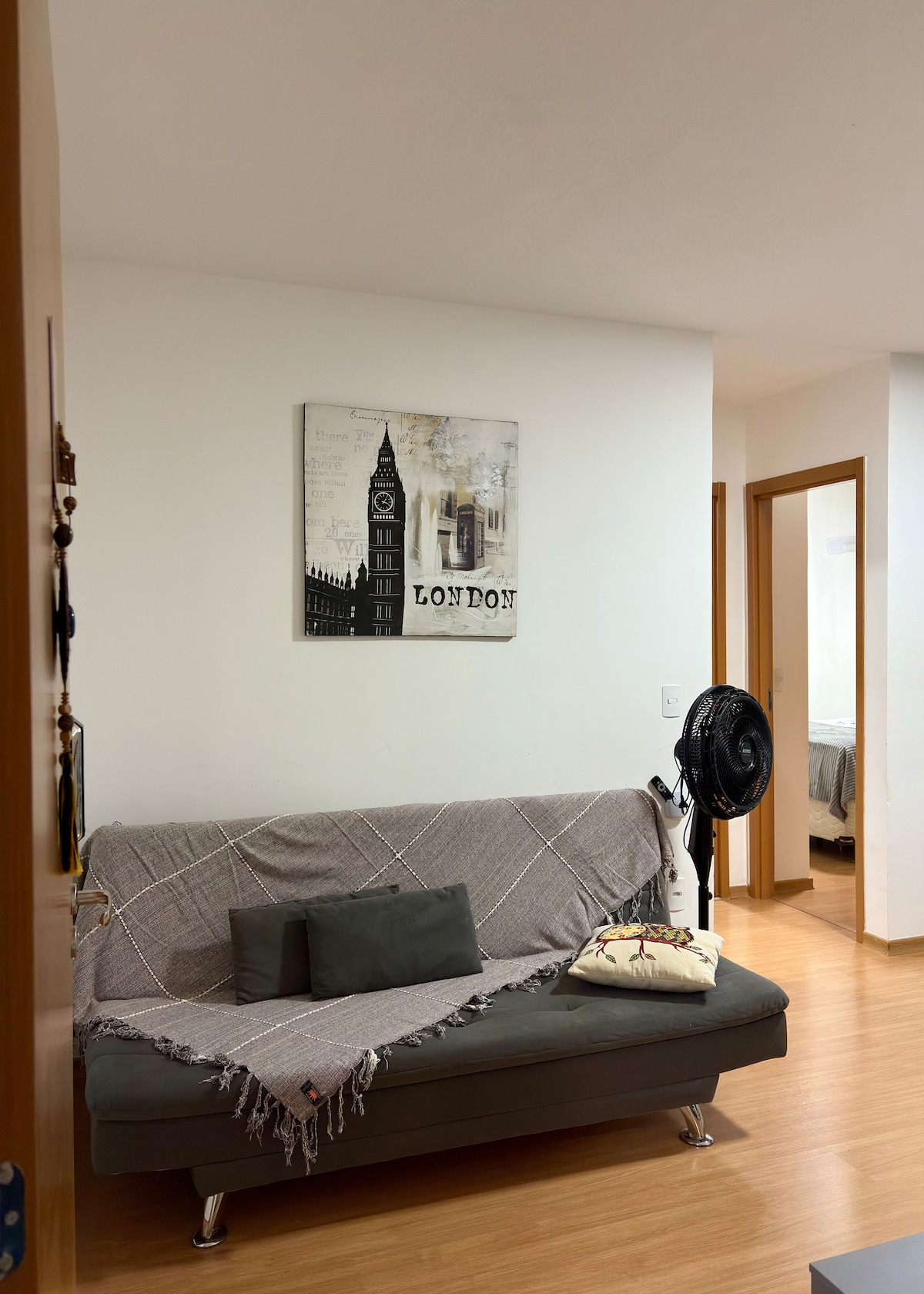 A cozy living area features a gray sofa bed adorned with a soft throw and decorative pillow. A wall art piece depicting a London clock tower is displayed above. A standing fan is positioned nearby, and doorways lead to other rooms in the background.