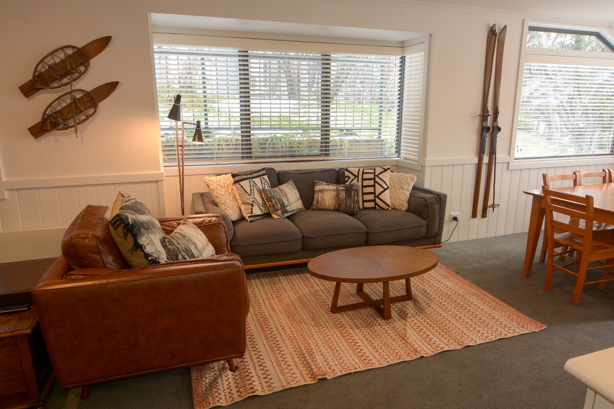 A welcoming living area features a comfortable gray sofa adorned with decorative pillows, complemented by a brown leather chair. A round wooden coffee table sits on a patterned rug, with a large window offering natural light. Decorative wooden skis and a lamp enhance the relaxed atmosphere.