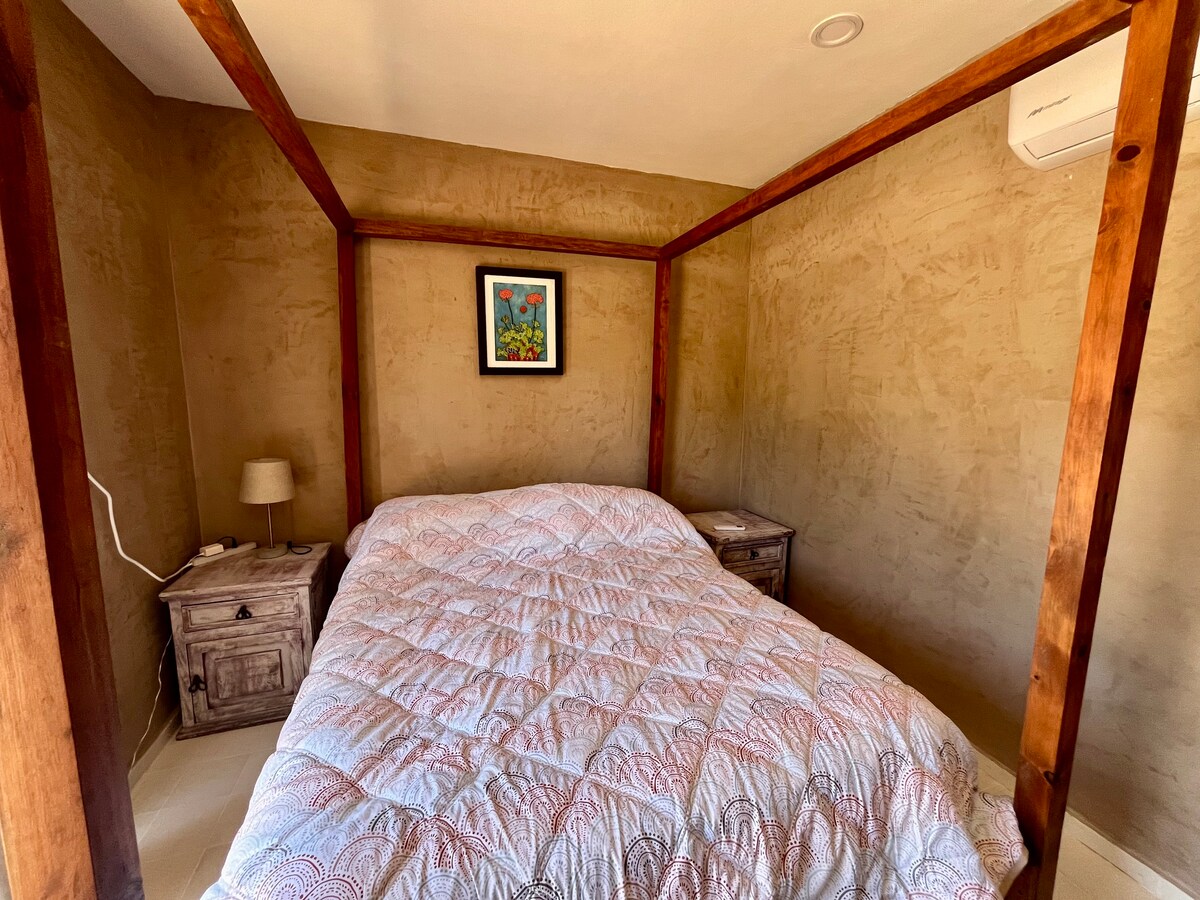A cozy sleeping area is furnished with a canopy bed, complemented by wooden nightstands on either side. A small lamp and framed artwork are present, with soft, neutral-toned walls providing a calming backdrop. Natural light filters into the space, creating a serene environment.