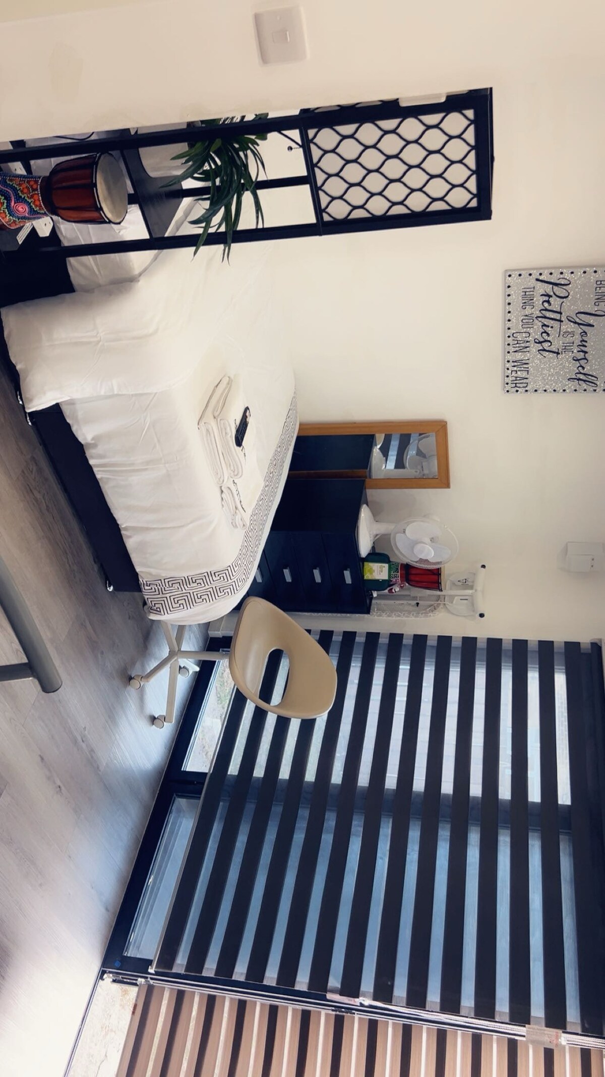 A cozy sleeping area is presented with a spacious bed adorned in neutral tones. A decorative screen adds a stylish touch, while a small chair and desk are positioned nearby. Natural light filters through dual-textured window coverings, enhancing the room’s relaxed ambiance.