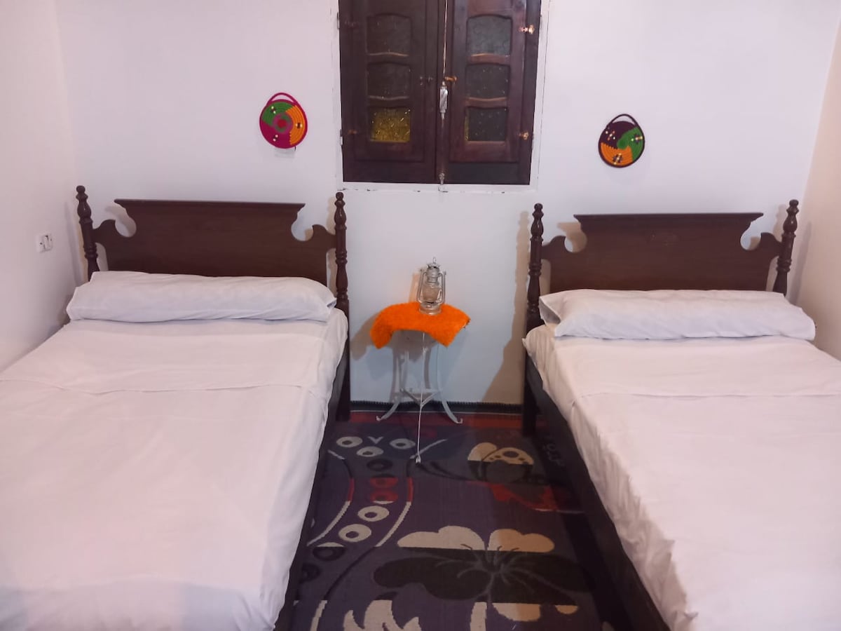A room features two spacious beds with crisp white linens, positioned parallel to each other. Colorful wall decorations add character to the room, and a small table with an orange cloth is centered between the beds. Natural light enters through a wooden window with intricate details.
