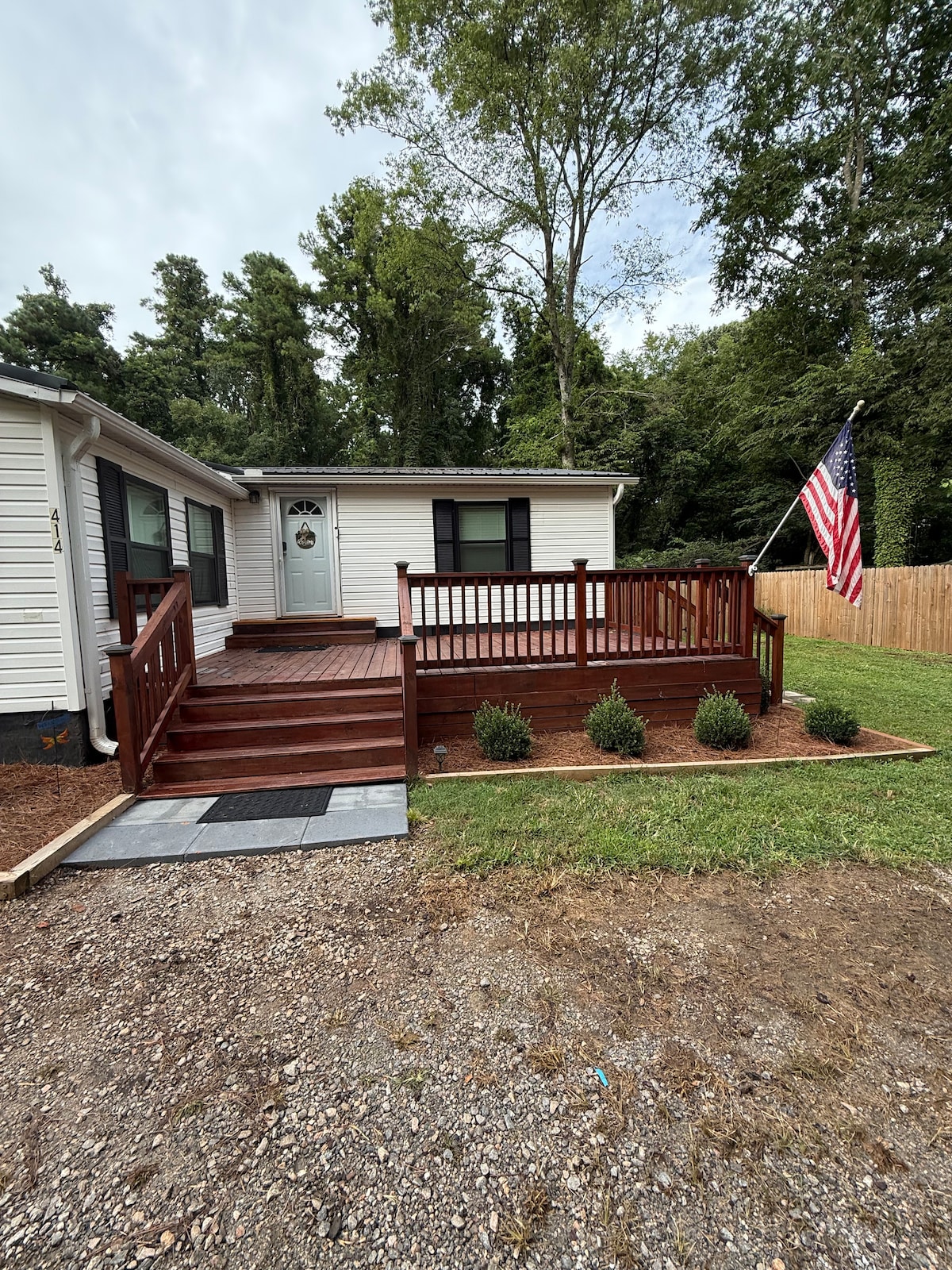 A charming exterior view of the home features a wooden deck with steps leading to the front door. An American flag is displayed on the side, complemented by neat landscaping with small shrubs. The surrounding area is grassy and bordered by a wooden fence.