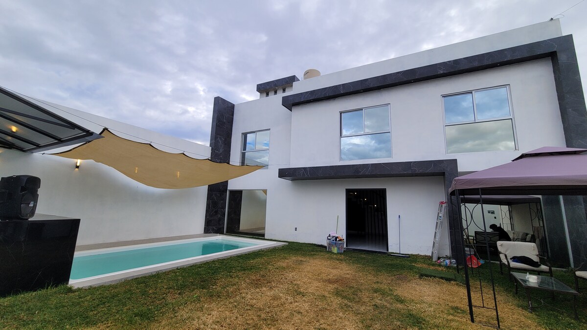 A contemporary two-story home is displayed, featuring large windows that reflect the cloudy sky. A sleek pool is positioned on the left, with a shaded area of fabric extending overhead. A modest lawn surrounds the pool area, enhancing the outdoor space.