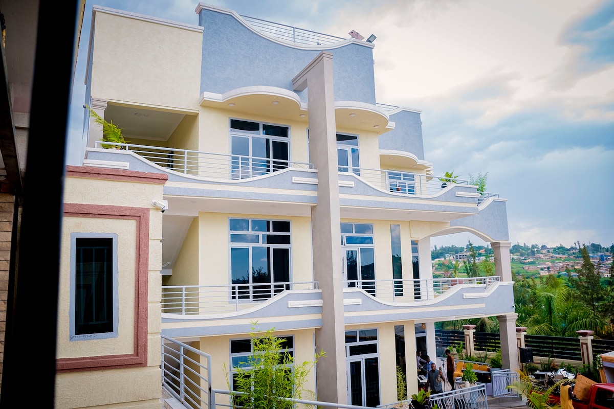 The exterior of a three-story apartment building is shown, featuring modern architectural lines and large glass windows. Balconies with railings extend from the second and third floors, and greenery is visible on the property, contributing to a serene environment.