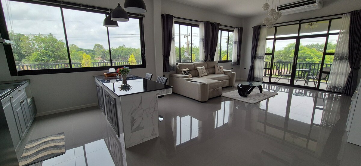 An open living area is paired with large windows, offering a view of lush greenery. A spacious sectional sofa faces a sleek coffee table, while a modern kitchen with a marble island is visible in the background. Light-colored walls and polished floors contribute to a bright atmosphere.