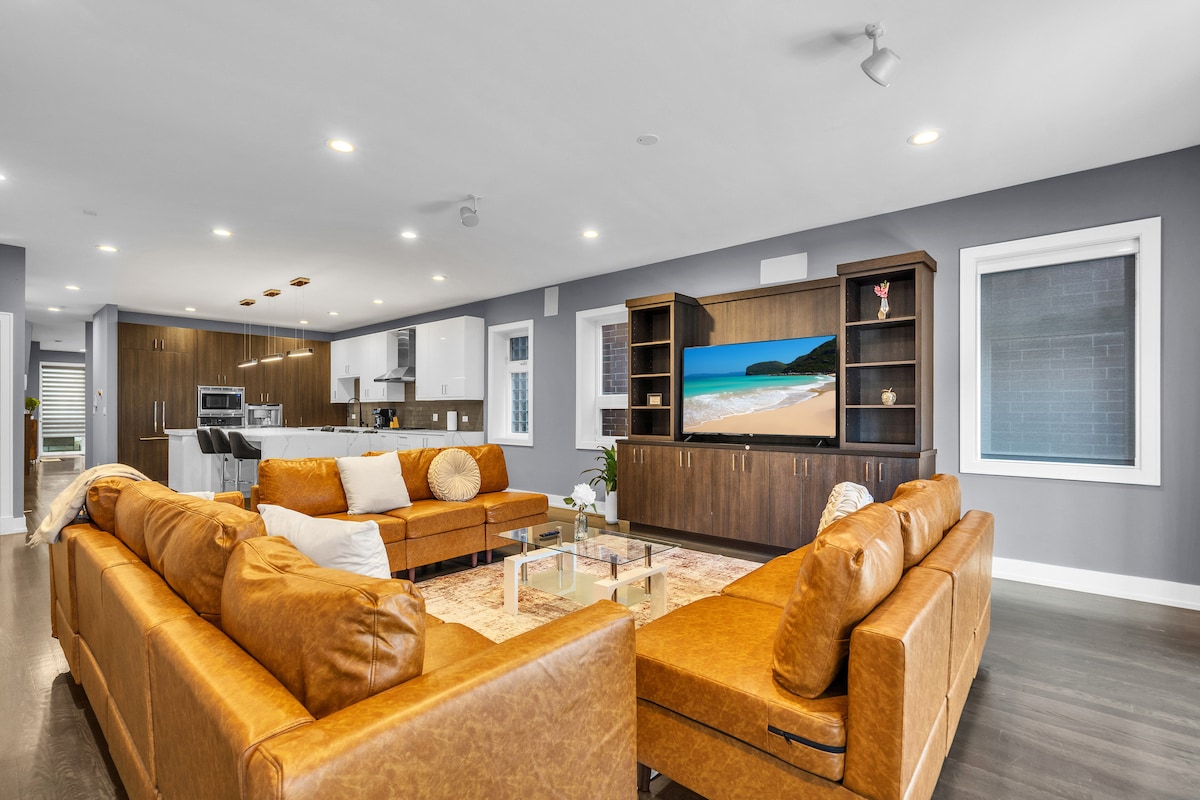A spacious living area is characterized by large, comfortable leather sofas arranged around a glass coffee table. A modern entertainment center with a television is visible, showcasing a serene beach scene. The room features hardwood floors and abundant natural light from windows.