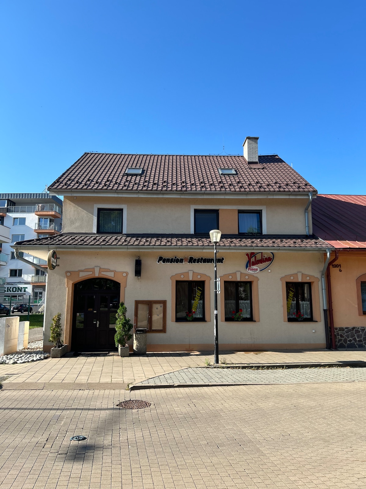 A quaint building features a charming façade with a mix of light and dark colors. Windows adorned with flower boxes add a touch of color. The entrance showcases a welcoming door, flanked by two small trees, while the surrounding area is paved with cobblestones.