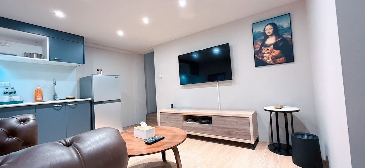 A modern living area features a large flat-screen television mounted on the wall. A sleek wooden media console sits below, and a round side table stands nearby. The kitchen area showcases cabinets and a refrigerator, contributing to the open space's functionality.