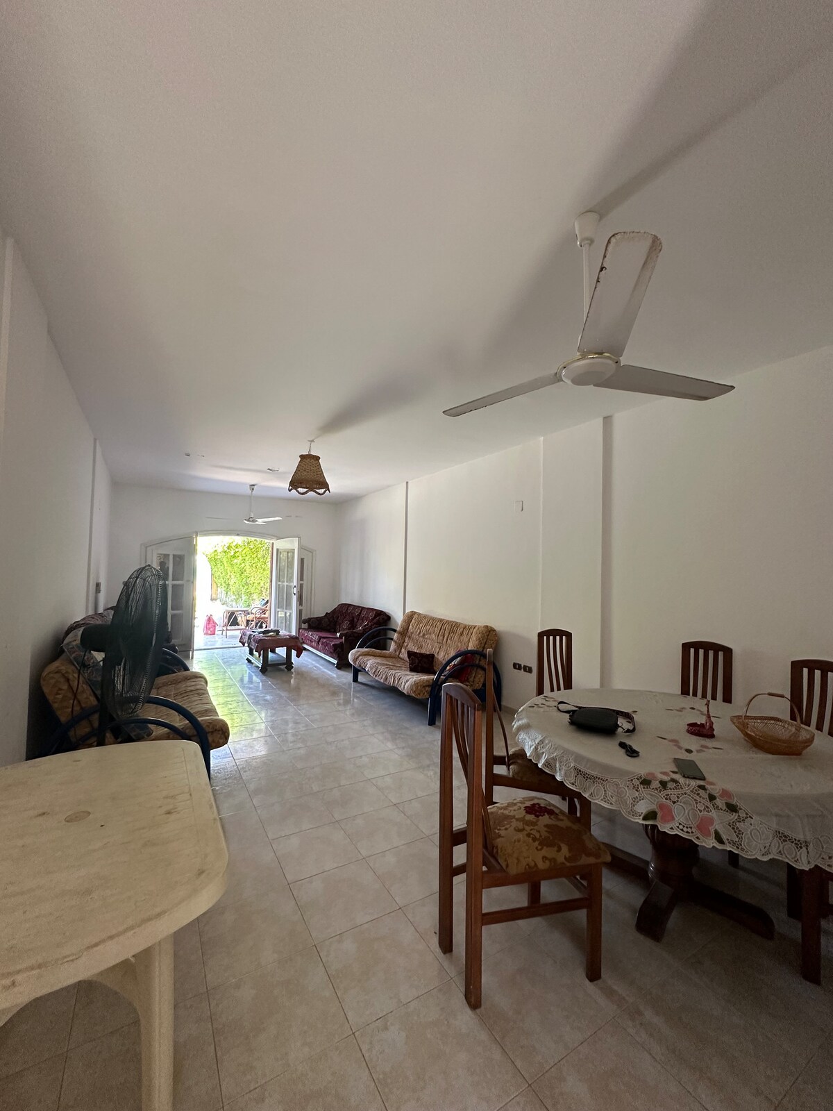 A spacious living area is designed with a combination of comfortable seating options, including a sofa and chairs. Natural light enters through the open double doors, leading to an outdoor space. A dining table is positioned nearby, surrounded by wooden chairs.