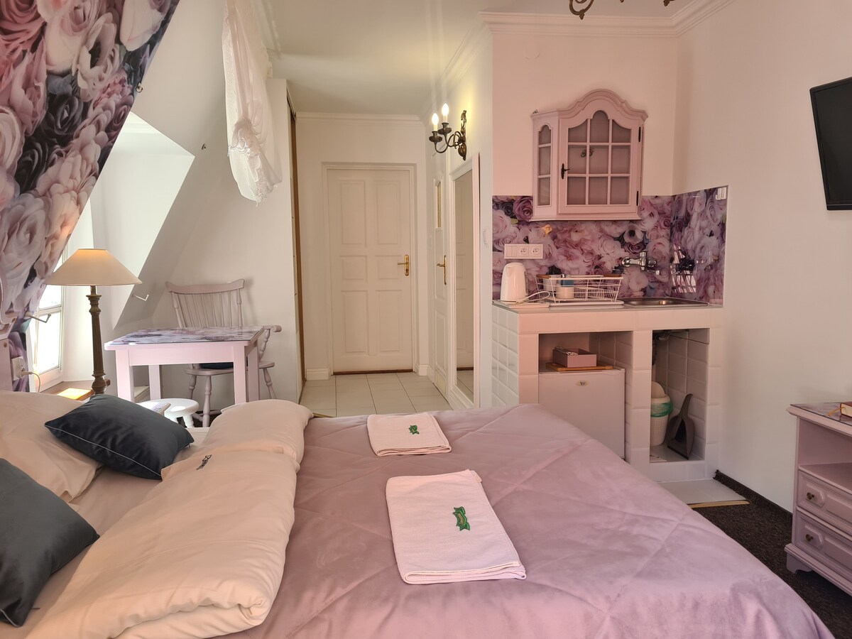 A well-appointed room features a spacious bed with soft linens and decorative pillows. A small kitchenette is visible, equipped with a sink and refrigerator. Floral wallpaper enhances the design. A dedicated workspace with a chair and table complements the welcoming atmosphere.