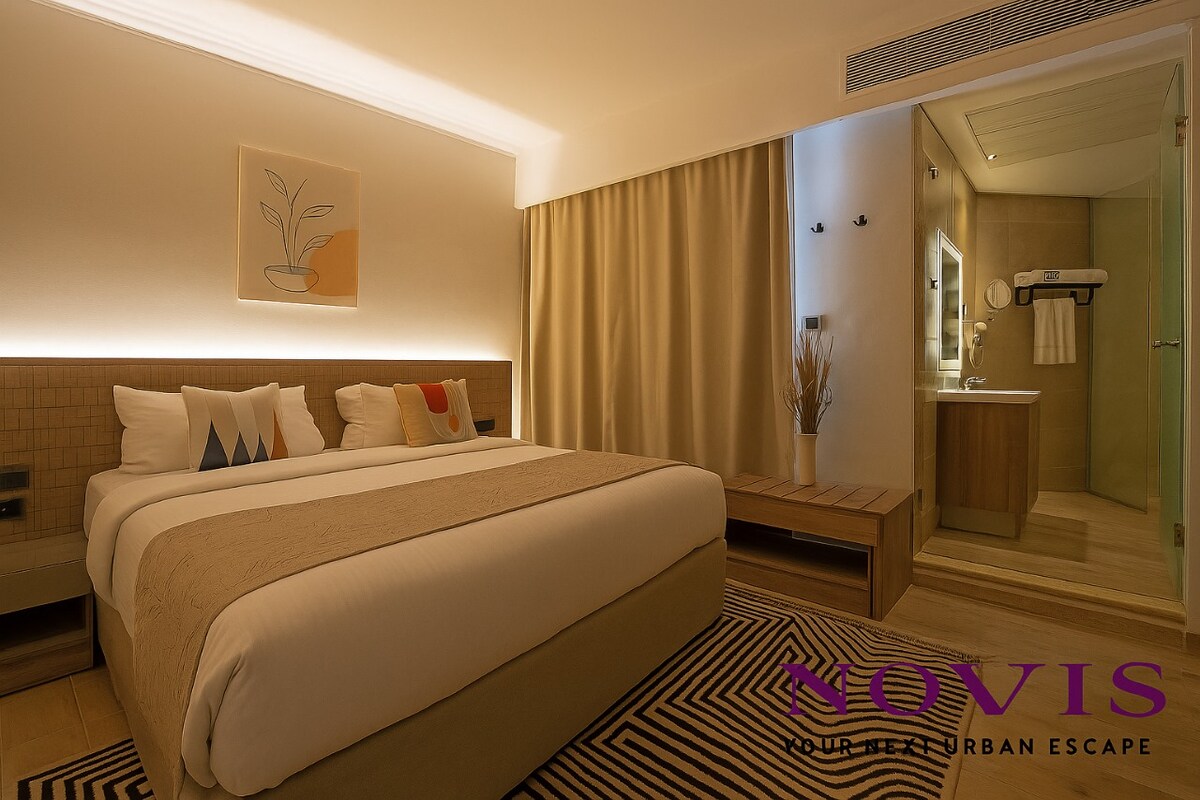 A contemporary bedroom features a spacious bed with neutral bedding and decorative pillows. Soft lighting accents the textured wall behind the bed. A seating area with a wooden bench is positioned next to large windows, while the en-suite bathroom is partially visible in the background.